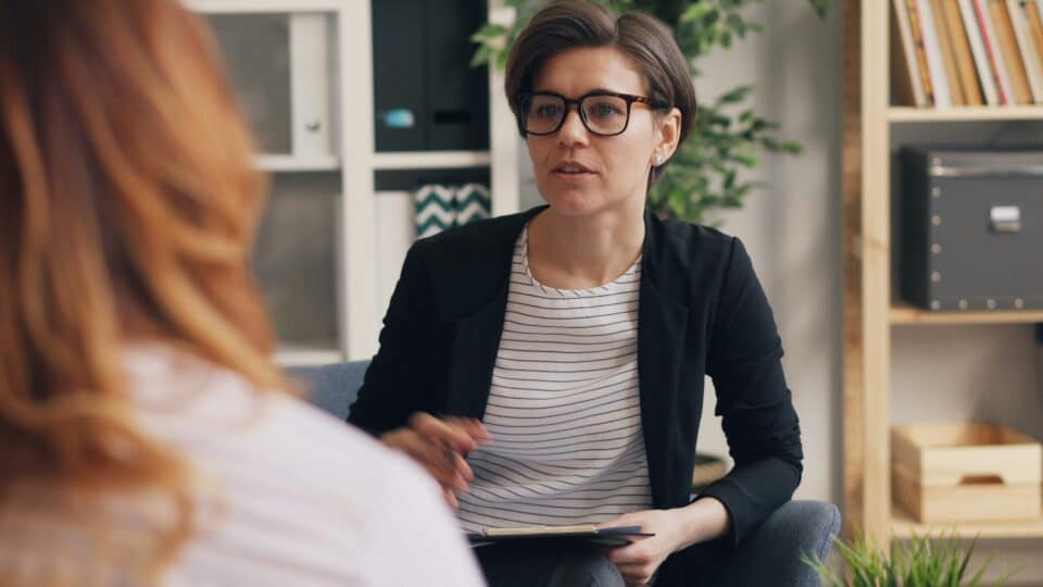 A woman in glasses engages in a conversation during a counseling session, seated in a stylish office with plants and shelves in the background.