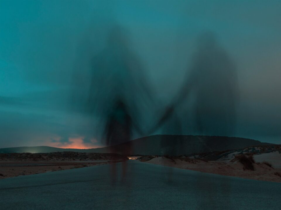 A blurred image of two figures holding hands on a deserted road under a moody sky at dusk.